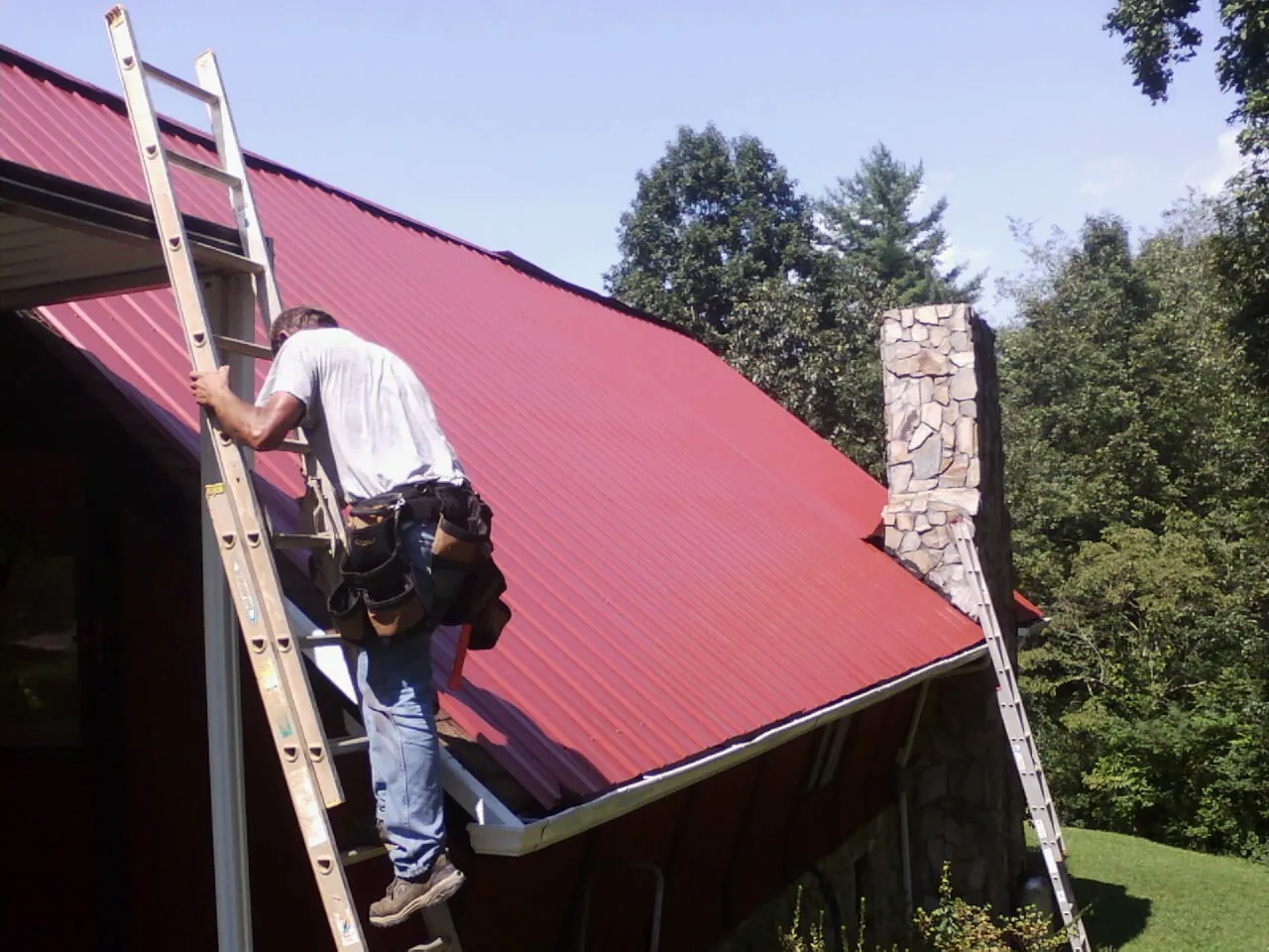 Roofer on a ladder beside a red metal roof and stone chimney — Soffit & Fascia Repair by Roofing Brothers in Grahamsville